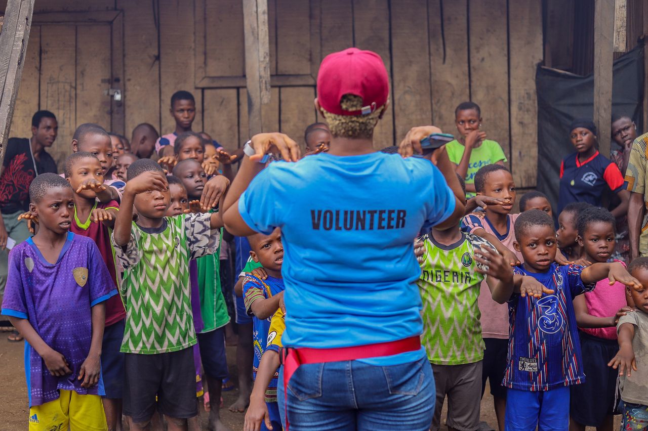 gallery-1 A volunteer guides a diverse group of children in an outdoor activity, fostering community spirit and teamwork.
